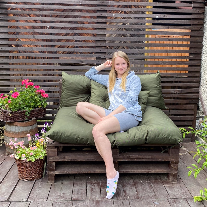 May include: A woman sits on a green cushioned pallet couch with a wooden frame. She is wearing a light blue hoodie and grey shorts. The couch is in front of a wooden slat wall.