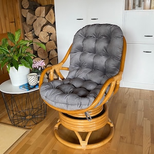 May include: A swivel rattan chair with a grey cushion. The chair is in a room with wood floors and a white cabinet in the background.