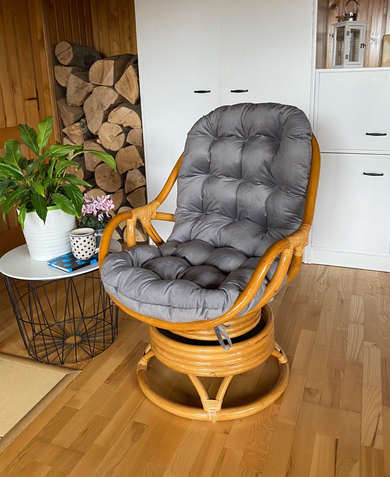 May include: A swivel rattan chair with a grey cushion. The chair is in a room with wood floors and a white cabinet in the background.