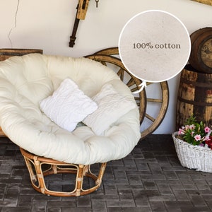 May include: A white, round, cushioned chair with a wicker frame and two white pillows. The chair is sitting on a brick patio. The chair is made of 100% cotton.