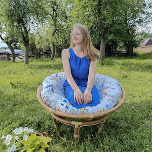 May include: A woman wearing a blue dress sits in a floral patterned chair with a wooden frame. The chair is on a grassy lawn.