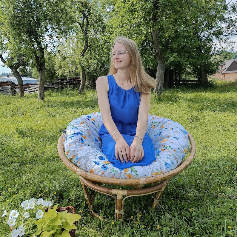May include: A woman wearing a blue dress sits in a floral patterned chair with a wooden frame. The chair is on a grassy lawn.