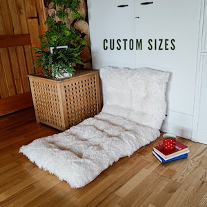May include: White fluffy floor cushion with a white pillow on top. The cushion is on a wooden floor with a wooden side table and a red and white mug on a stack of books. The text "CUSTOM SIZES" is visible on a white cabinet in the background.