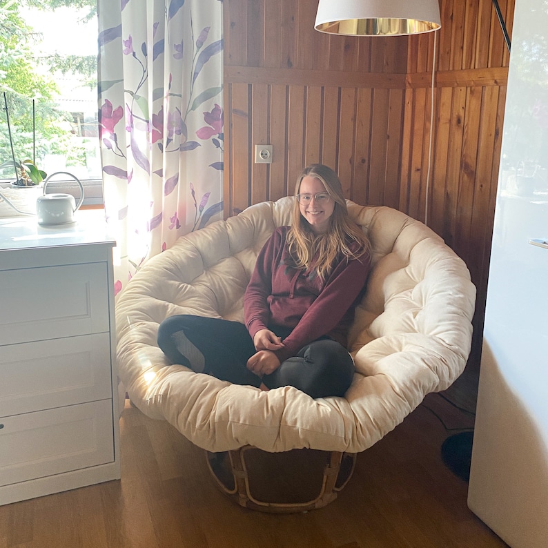 May include: A woman sits in a large, white, round, cushioned chair with a wooden base. The chair is in a room with wood paneling on the walls and a window with white curtains.