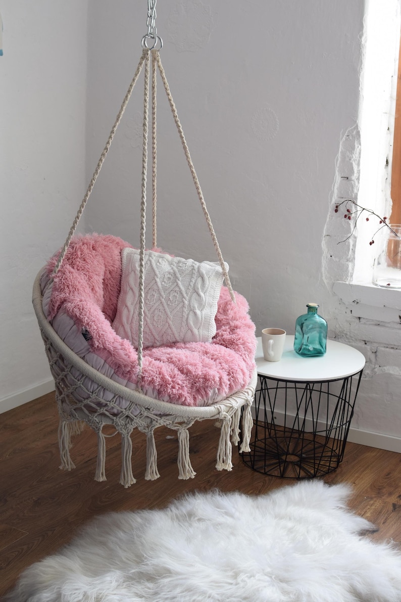 Puede incluir: Una silla colgante rosa y blanca con una almohada de punto blanco. La silla est&aacute; suspendida del techo con una cuerda y tiene una franja alrededor de la parte inferior.