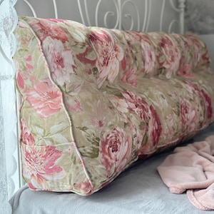 May include: A floral-patterned bed rest pillow with a beige background and pink flowers. The pillow is rectangular with a curved top and a white trim. It is placed on a bed with a light blue sheet and a pink blanket.
