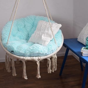 Puede incluir: Una silla colgante de color azul claro con una almohada de punto blanco y detalles de flecos. La silla est&aacute; colgada de un techo blanco y est&aacute; frente a una pared blanca.