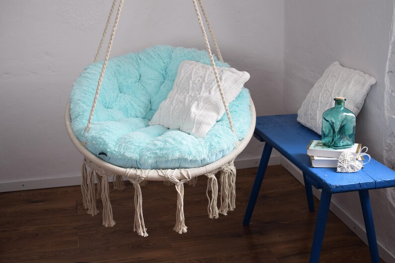 Puede incluir: Una silla colgante de color azul claro con una almohada de punto blanco y detalles de flecos. La silla est&aacute; colgada de un techo blanco y est&aacute; frente a una pared blanca.