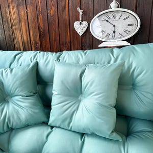 May include: A teal-colored couch with three cushions. The couch is against a wooden wall with a white clock and a white heart hanging on the wall.