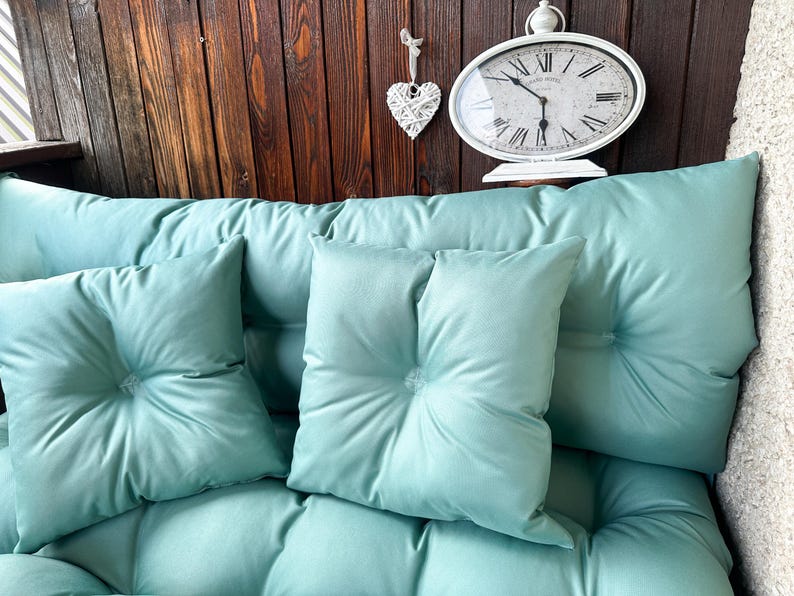 May include: A teal-colored couch with three cushions. The couch is against a wooden wall with a white clock and a white heart hanging on the wall.