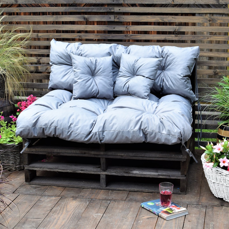 May include: A gray pallet couch with a gray cushion and four gray throw pillows. The couch is on a wooden deck with a book and a glass of red liquid on the deck.