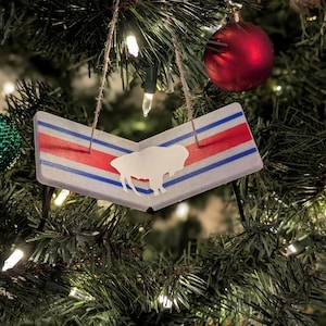 May include: A Christmas tree ornament featuring a white bison silhouette on a rectangular design with red and blue stripes. The ornament hangs from a twine string and is surrounded by Christmas tree branches, lights, and ornaments.