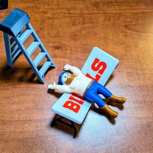 Pode incluir: Uma pequena figura de plástico de uma pessoa barbuda com capacete azul, calças azuis e botas castanhas, deitada numa mesa dobrável branca com o texto vermelho "BILLS". Ao lado da mesa encontra-se um pequeno escadote de plástico azul-claro.