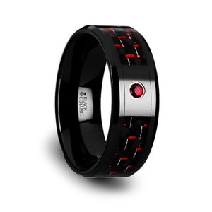 May include: Black ceramic ring with a red and black carbon fiber inlay. The ring features a small, round, red gemstone set in a silver-colored metal band. The text "BLACK CERAMIC" is visible on the side of the ring.