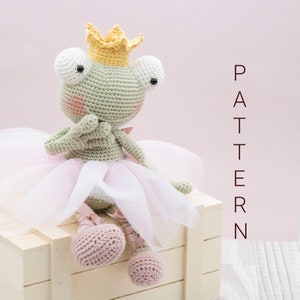 Puede incluir: Una muñeca de rana de ganchillo con una corona amarilla y un tutú rosa. La rana es de color verde claro con ojos blancos y mejillas rosas. La muñeca está sentada en una caja de madera blanca. La palabra "PATTERN" está escrita verticalmente en el fondo.