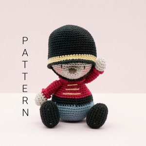 May include: A handmade crochet doll resembling a British Royal Guard, with a black hat, red jacket, and blue and black trousers. The word "PATTERN" is written vertically on the left side.