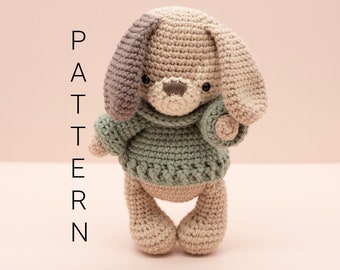 May include: A crocheted stuffed dog wearing a green sweater. The dog has brown and gray fur and black eyes. The dog is standing on a pink surface.