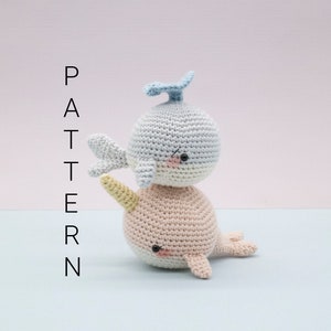 May include: A crocheted narwhal toy with a light blue body and a yellow horn, stacked on top of a crocheted whale toy with a light pink body and a blue fin. Both toys have black eyes and a stitched smile.