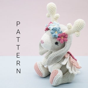 May include: A crocheted stuffed animal resembling a deer with a floral crown. The deer is light grey with pink accents and has large, floppy ears. The floral crown is made of blue and pink flowers.