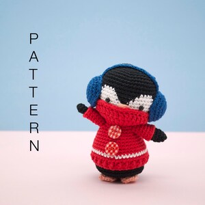Puede incluir: Un pingüino de crochet que lleva un suéter rojo y blanco y orejeras azules. El pingüino tiene rasgos faciales negros y blancos y botones rojos en su suéter.