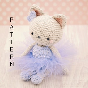 May include: A crocheted cat wearing a light blue tutu with a pink flower on its head. The cat is sitting on a light wood surface.