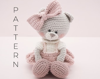 May include: A crocheted teddy bear wearing a pink dress and a pink bow. The bear has grey fur and black eyes.