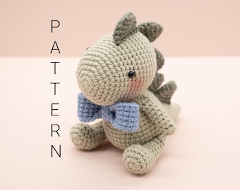May include: A crocheted dinosaur toy with a light green body, a blue bow tie, and black eyes. The toy is sitting on a pink surface.
