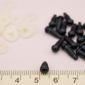 Może przedstawiać: Czarne plastikowe oczka bezpieczeństwa z gwintowanym tyłem, o średnicy około 8 mm. Oczka są pokazane obok linijki z liczbami 4, 5, 6 i 7.