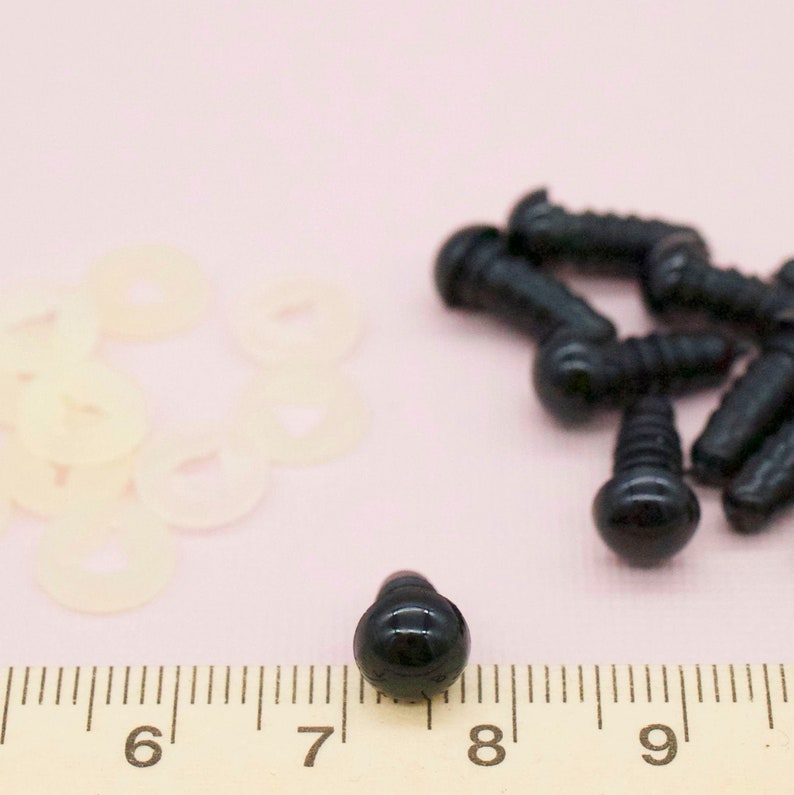 K&ouml;nnte beinhalten: Schwarze Kunststoff-Sicherheitsaugen f&uuml;r Bastelarbeiten, mit einem Durchmesser von 2,2 cm. Die Augen sind auf einem Lineal mit einem Stapel wei&szlig;er Kunststoffscheiben abgebildet.