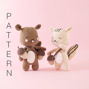 May include: Two crocheted squirrel amigurumi toys, one brown and one beige, holding acorns. The squirrels have black eyes and smiling mouths. The toys are on a pink background.