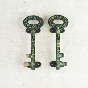 May include: Two decorative antique-style keys. The keys are black with green floral designs and brass-coloured accents. The oval-shaped key heads have a decorative pattern. The keys are mounted on a light-coloured surface.