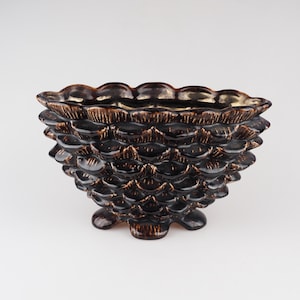 May include: A dark brown ceramic bowl with a pine cone design. The bowl has a scalloped rim and three feet.