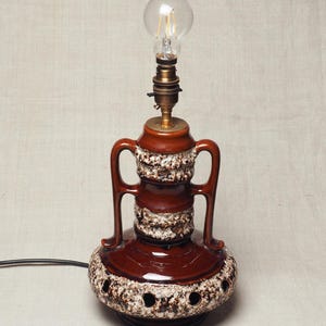 May include: A ceramic table lamp featuring a glossy brown glaze with textured white and dark brown lava-style accents. The lamp has two side handles, a wide base with circular cutouts, a brass-coloured socket, and a clear glass LED light bulb.
