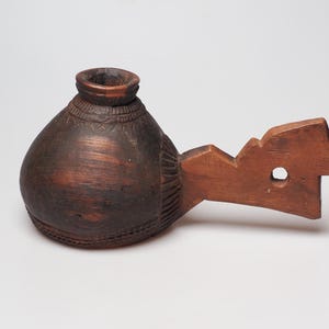 May include: A hand-carved wooden vessel with a bulbous body and a handle. The vessel is dark brown with lighter brown accents, and has decorative carvings around the top and base. The handle has a geometric design with a circular hole.