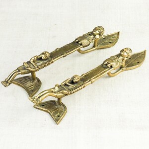 May include: A pair of antique brass door handles. Each handle has a detailed design of a reclining figure on a textured bar, with a leaf-shaped base. The handles are a warm gold colour, adding a vintage touch to any door.