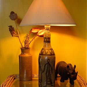 May include: A table lamp with a beige conical shade and a ceramic bottle base featuring hand-painted dark brown abstract figures and geometric patterns. Beside it sits a plain cylindrical ceramic vase with dried stems and a black rhinoceros figurine.