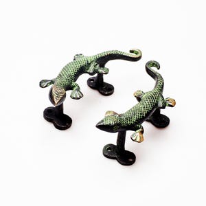 May include: Three bronze-coloured lizard-shaped cabinet pulls. The lizards have a textured, scale-like pattern in green and gold. Each lizard is mounted on a black base with a single screw hole. The pulls are arranged on a white background.