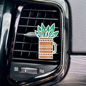 May include: A green plant air freshener in a brown wooden pot hanging from a car's air vent.