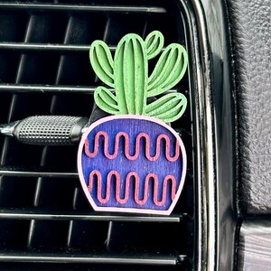May include: A green and purple wooden car air freshener shaped like a potted plant. The plant has a green stem and leaves. The pot is purple with pink wavy lines.