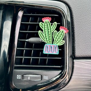 May include: A green cactus air freshener with pink flowers in a blue and pink pot. The air freshener is hanging from a car's air vent.