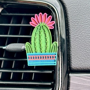 May include: A green and pink wooden cactus air freshener with a blue and pink pot. The cactus is in a car's air vent.