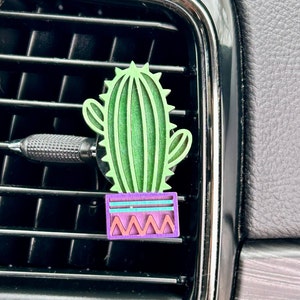 May include: A green and purple cactus air freshener hanging from a car's air vent. The cactus is in a small pot with a geometric pattern.