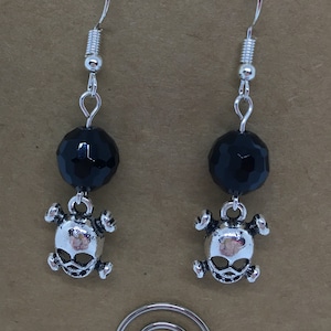 May include: A pair of silver skull and black crystal earrings. The earrings are on a brown card with the text "Ginger Constantino for GINGERBREAD HEARTH".