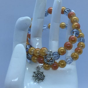 Könnte beinhalten: Zwei Perlenarmbänder mit orangefarbenen, pfirsichfarbenen und klaren Perlen. Die Armbänder haben einen silbernen Kürbis-Anhänger und einen silbernen Spinnennetz-Anhänger.