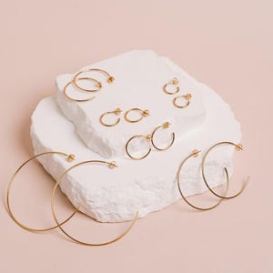 Peut inclure: Une collection de boucles d'oreilles créoles dorées de différentes tailles, présentées sur une surface blanche texturée sur fond rose clair. Les boucles d'oreilles vont des petits clous aux grandes créoles circulaires.