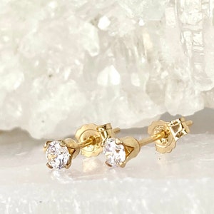 May include: A pair of gold stud earrings with clear, round gemstones. The earrings are set in a classic four-prong setting, and the posts have butterfly backs. The earrings are displayed on a white surface.