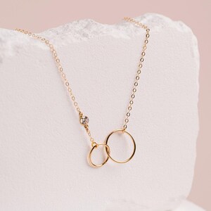 A delicate gold necklace with two smooth linked circles, one larger and one smaller, is displayed against a light pink background.