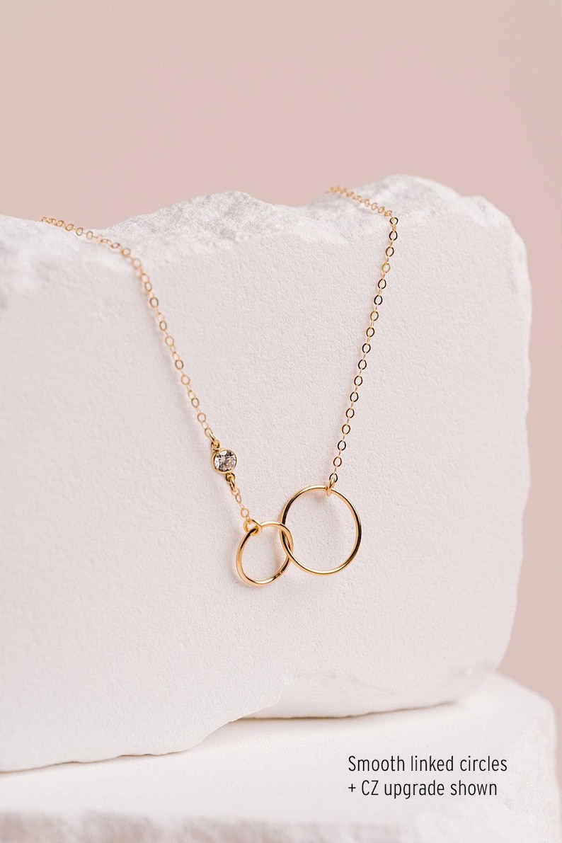 A delicate gold necklace with two smooth linked circles, one larger and one smaller, is displayed against a light pink background.