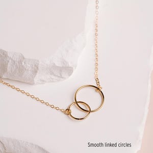 A delicate gold necklace with two circular pendants, one larger and one smaller, is displayed on a white surface.