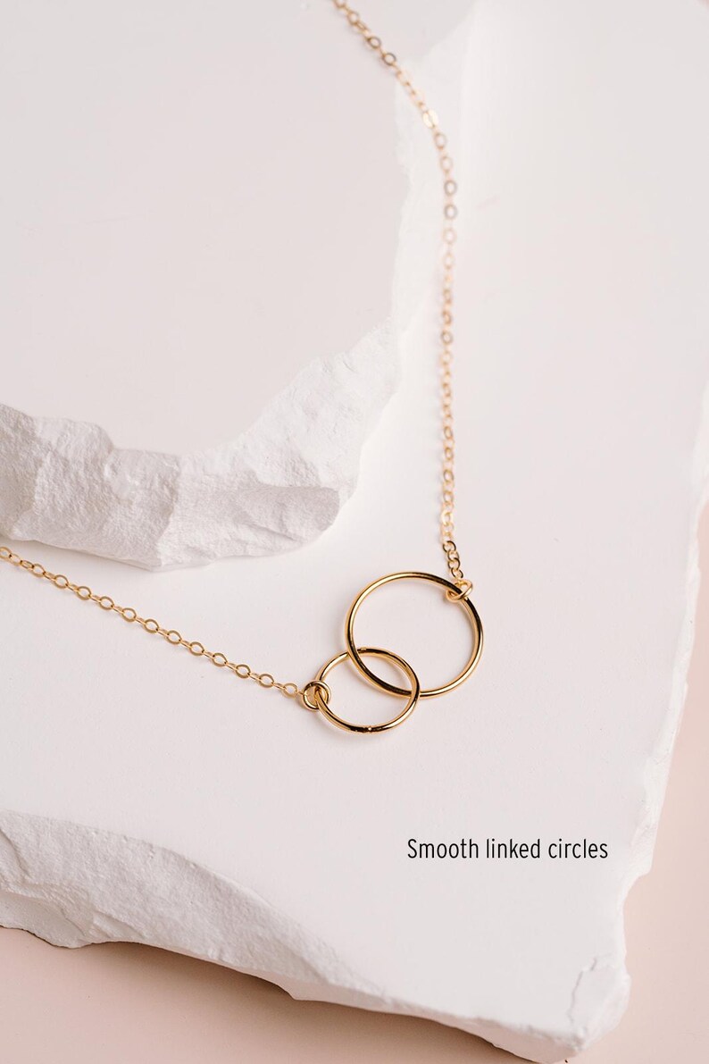 A delicate gold necklace with two circular pendants, one larger and one smaller, is displayed on a white surface.